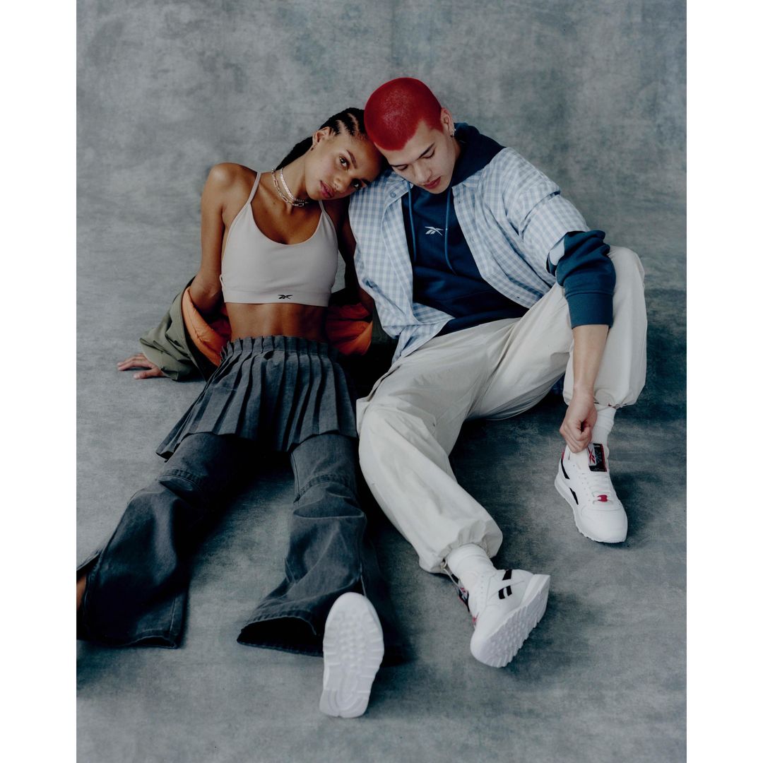 An adult woman and an adult man, posing on the floor with their heads touching and wearing athleisure wear in a photo studio setting.