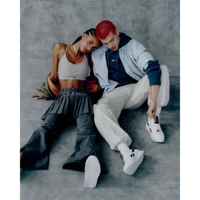 An adult woman and an adult man, posing on the floor with their heads touching and wearing athleisure wear in a photo studio setting.