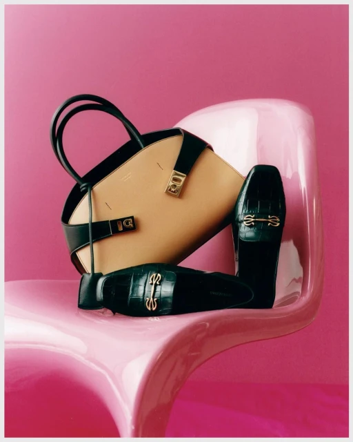 Fashion still life of a beige handbag with black straps and black loafers with gold hardware, staged on a pink plastic chair.