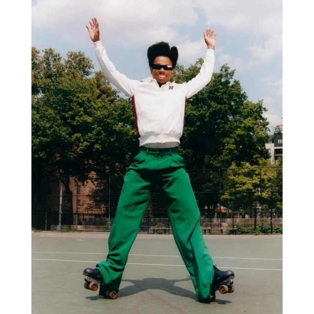 Adult roller skater in bright green pants and a white jacket jumps in the air with arms raised against an outdoor court and trees.