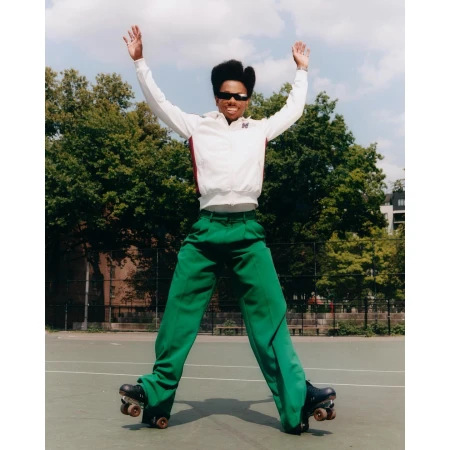 Adult roller skater in bright green pants and a white jacket jumps in the air with arms raised against an outdoor court and trees.