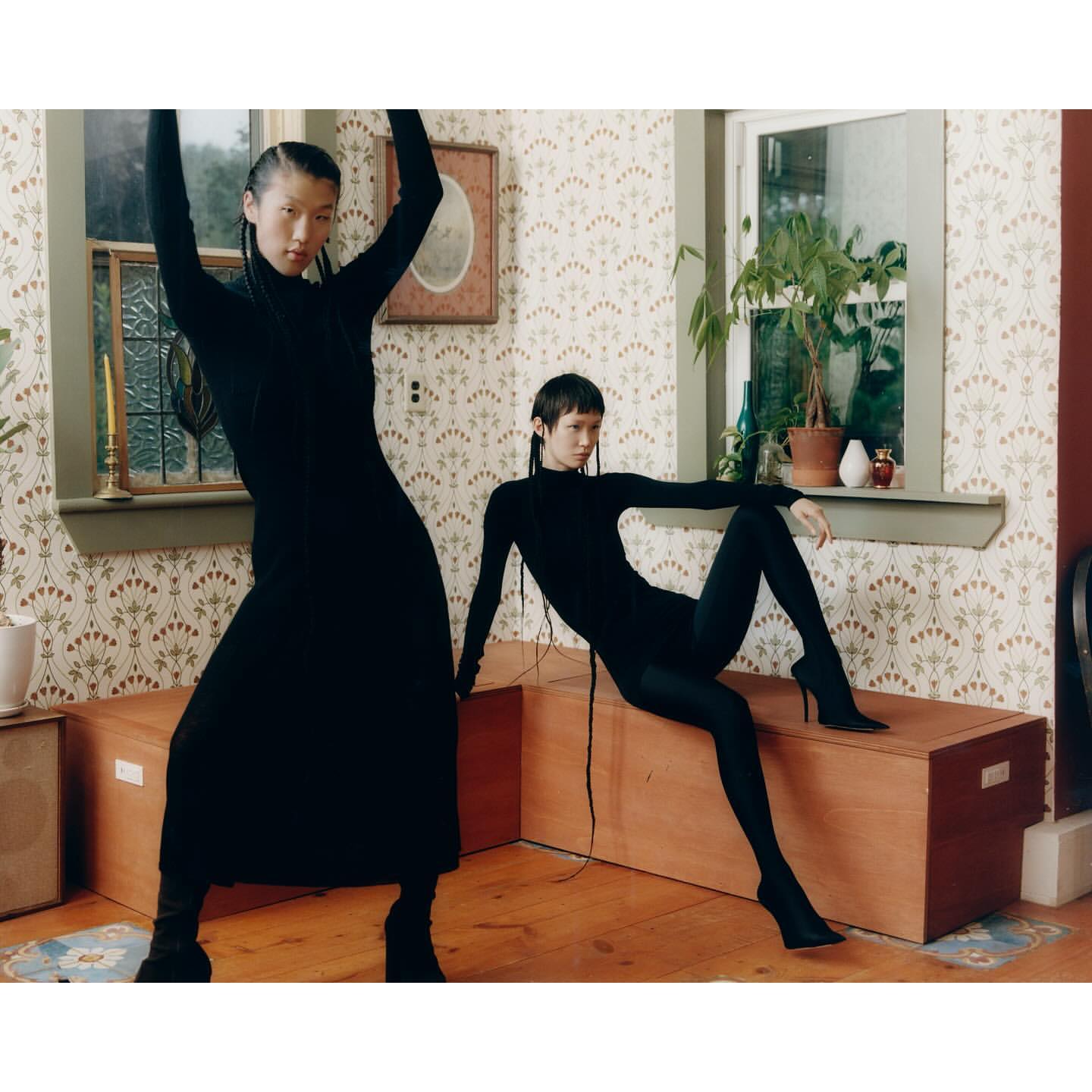 Two women pose in long-sleeved black dresses and boots inside a room with patterned wallpaper, a window, and a wooden chest.