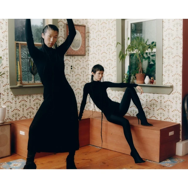 Two women pose in long-sleeved black dresses and boots inside a room with patterned wallpaper, a window, and a wooden chest.