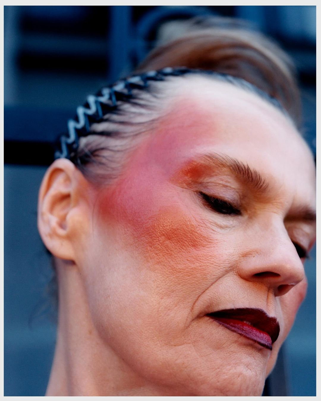 Close-up portrait of an older woman with bold red blush on her cheek and dark lipstick, wearing a black headband.
