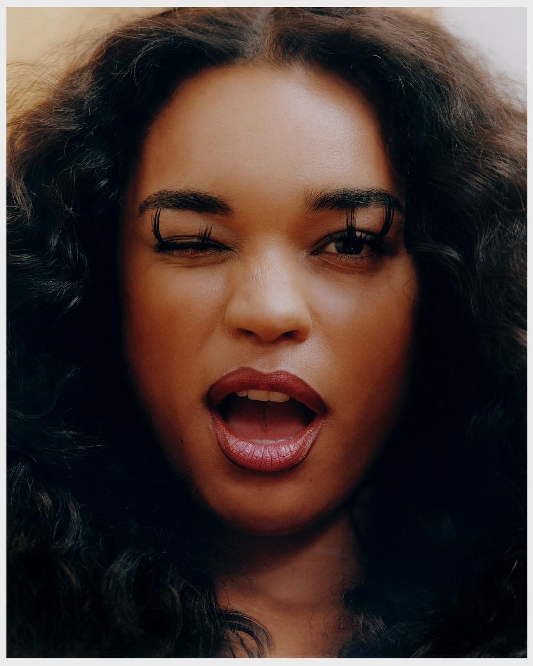 Close-up portrait of a woman with dark, wavy hair, winking with long lashes, and a partially open mouth, set against a neutral background.