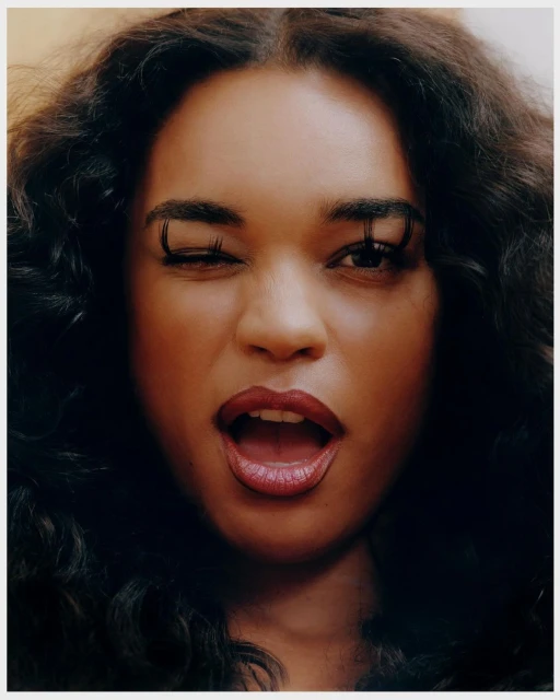 Close-up portrait of a woman with dark, wavy hair, winking with long lashes, and a partially open mouth, set against a neutral background.