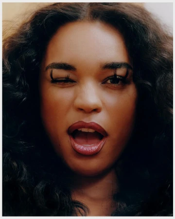 Close-up portrait of a woman with dark, wavy hair, winking with long lashes, and a partially open mouth, set against a neutral background.