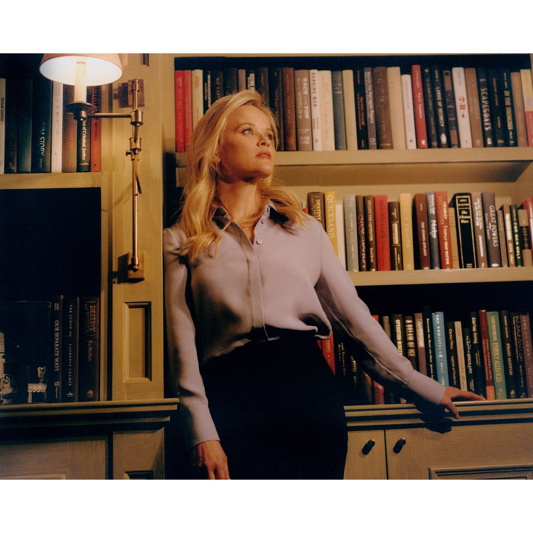 Portrait of a blonde woman in a lilac collared shirt leaning against a bookcase indoors, illuminated by a nearby lamp.