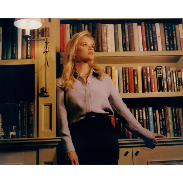 Portrait of a blonde woman in a lilac collared shirt leaning against a bookcase indoors, illuminated by a nearby lamp.