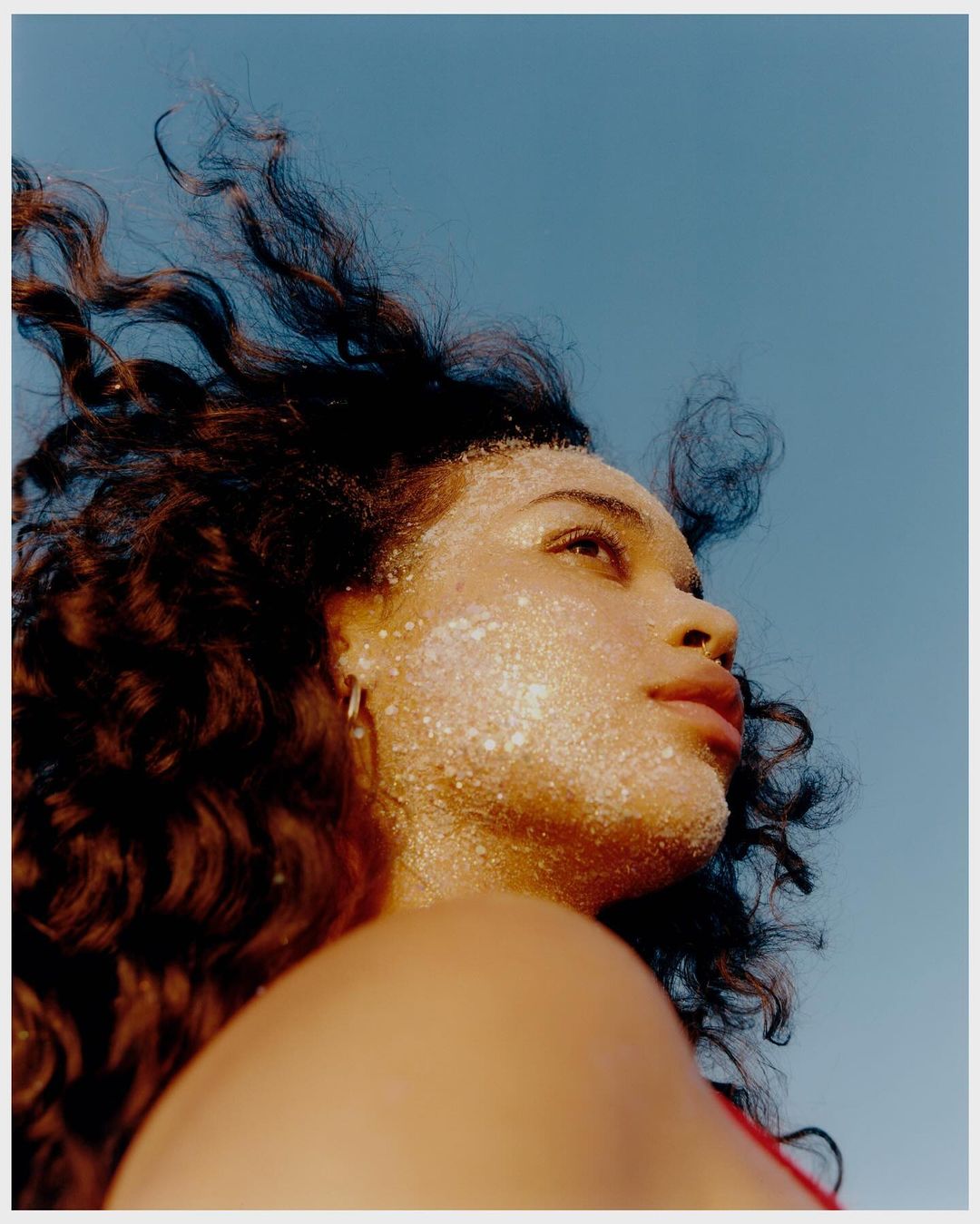 Portrait of a woman with glitter on her face and curly hair, looking up against a clear blue sky in an outdoor setting.