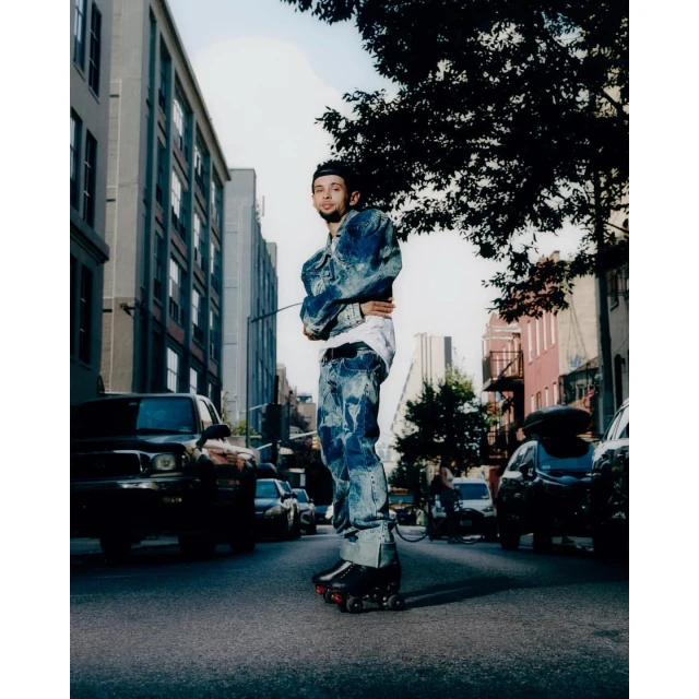Man in a patterned denim jacket and pants standing on asphalt street wearing roller skates, city buildings and cars in the background.