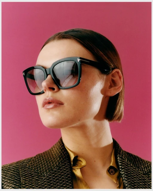 Close-up shot of a stylish adult woman wearing black, square-framed sunglasses and a textured brown jacket against a solid pink backdrop.