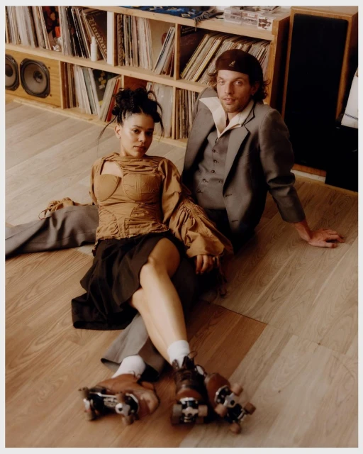 Woman on roller skates poses with a man in a suit near a record collection, indoor setting with natural light and a wooden floor.