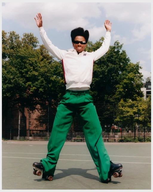 Person on roller skates jumps with arms up in the air on an outdoor sports court with trees, buildings, and sky in the background.