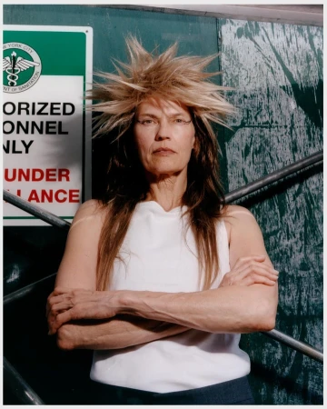 Portrait of an adult woman with spiked blonde hair standing in front of a green sanitation sign and metal bars with arms crossed.