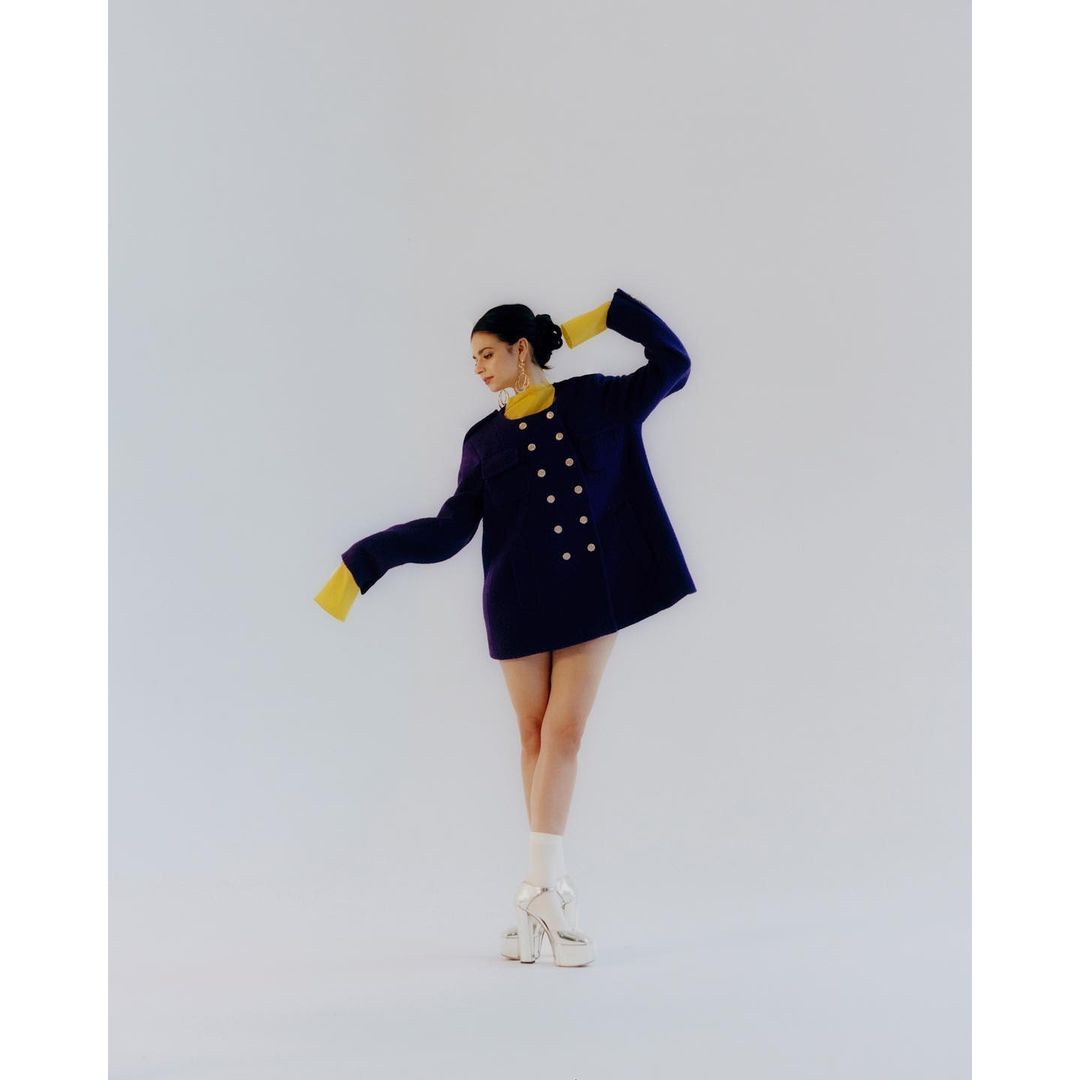 Woman in navy coat with gold buttons and yellow sleeves poses indoors in white heels and socks for a fashion forward studio shot.