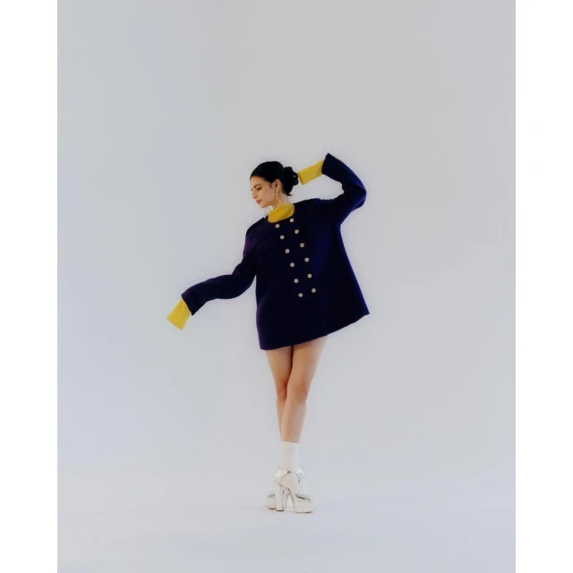 Woman in navy coat with gold buttons and yellow sleeves poses indoors in white heels and socks for a fashion forward studio shot.