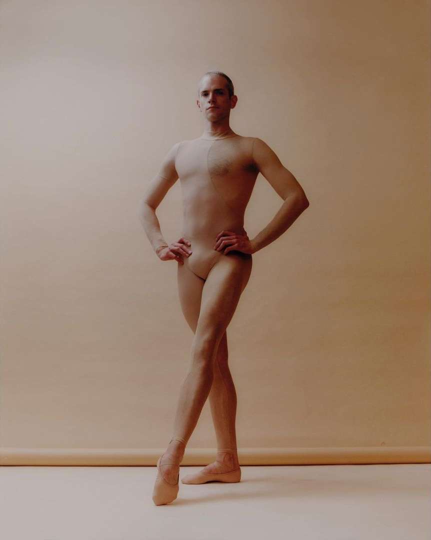 A male ballet dancer poses in a studio, wearing flesh-colored clothing and pointe shoes, against a light beige background. Full-body shot.