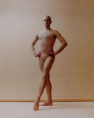 A male ballet dancer poses in a studio, wearing flesh-colored clothing and pointe shoes, against a light beige background. Full-body shot.