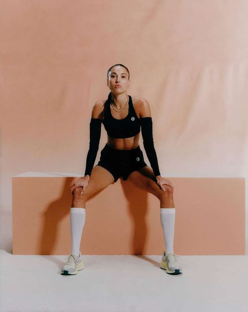 Athletic woman in workout attire rests with hands on knees in a studio, showcasing sport and fitness lifestyle against a light orange background.