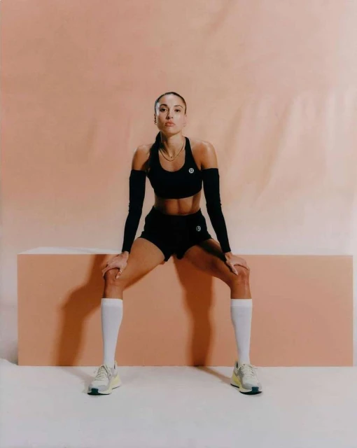 Athletic woman in workout attire rests with hands on knees in a studio, showcasing sport and fitness lifestyle against a light orange background.