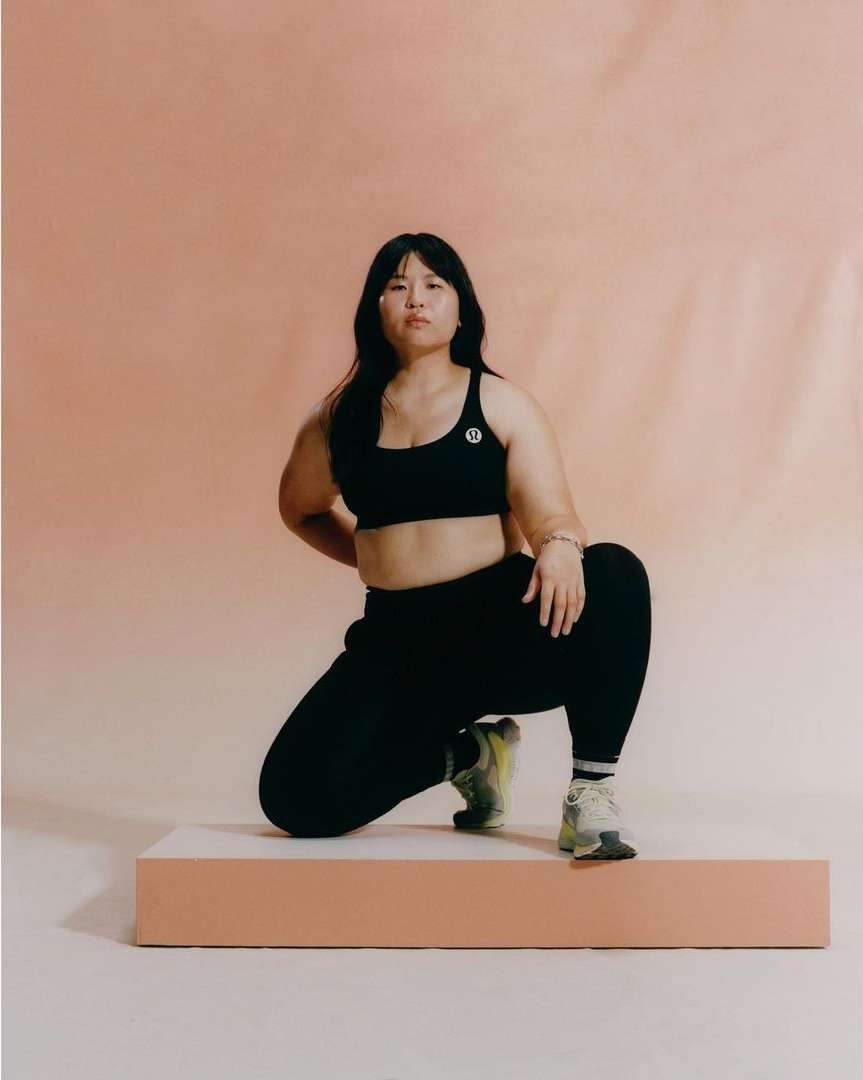 East Asian woman in black athletic wear kneeling on a low peach-colored platform against a warm neutral backdrop