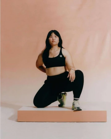 East Asian woman in black athletic wear kneeling on a low peach-colored platform against a warm neutral backdrop
