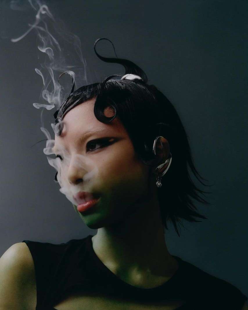 Portrait of an Asian woman with dramatic eye makeup and sculpted black hair, partially obscured by smoke, set against a dark backdrop.