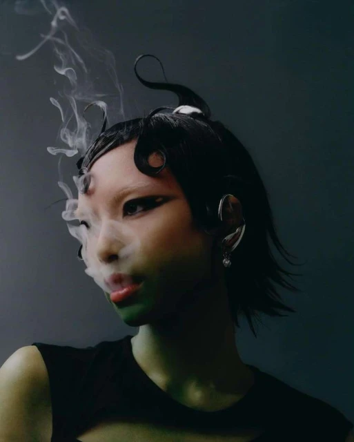 Portrait of an Asian woman with dramatic eye makeup and sculpted black hair, partially obscured by smoke, set against a dark backdrop.