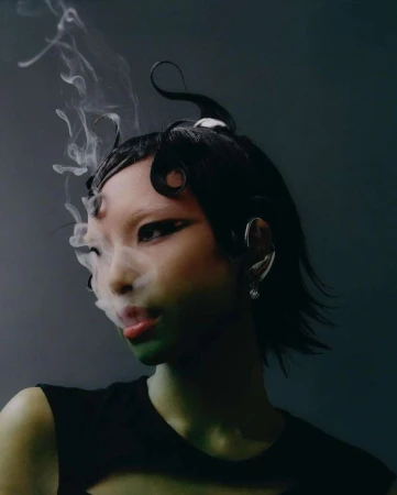 Portrait of an Asian woman with dramatic eye makeup and sculpted black hair, partially obscured by smoke, set against a dark backdrop.