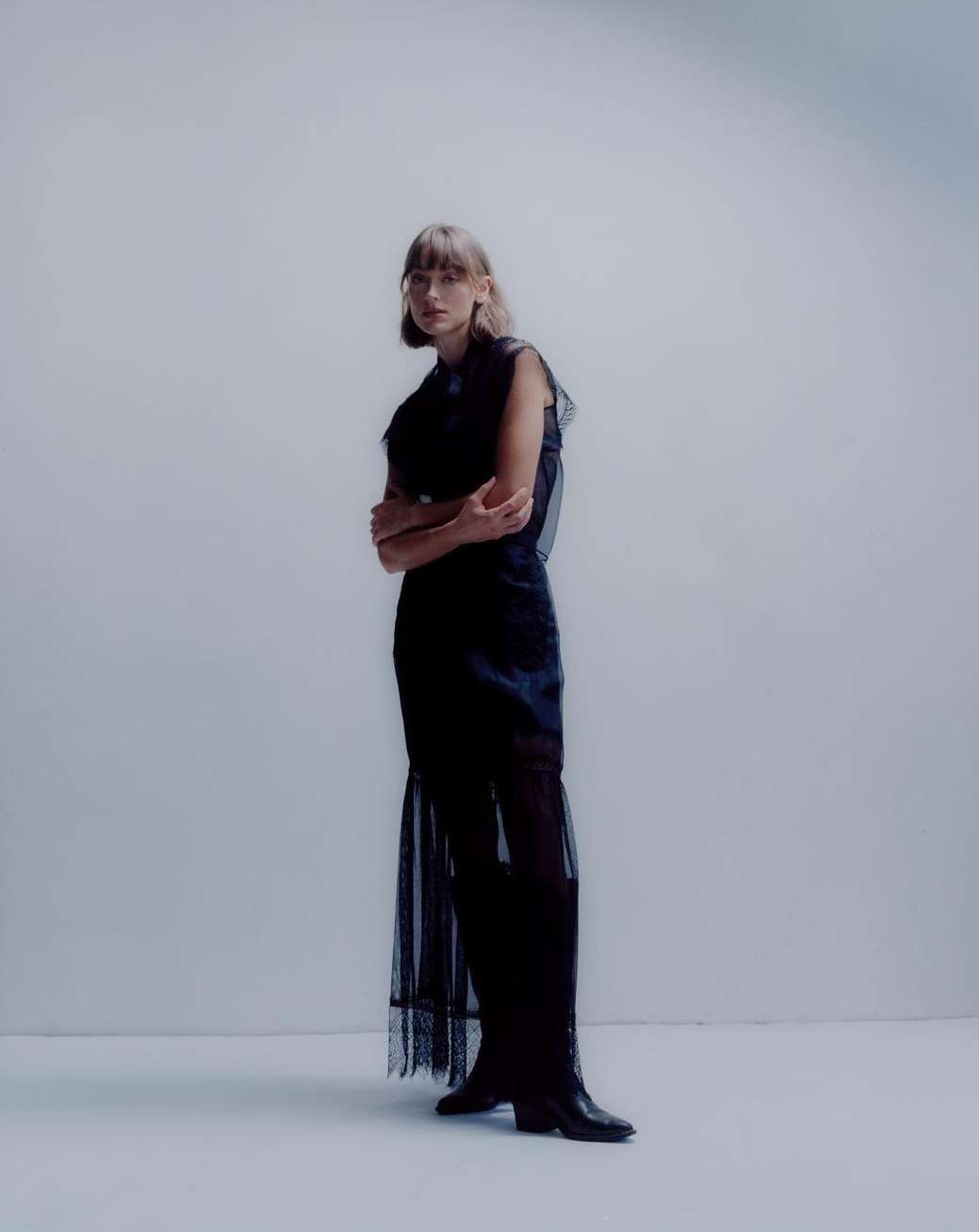 Fashion model in a long dark dress stands in a photo studio with arms crossed, posing for a fashion shot in a studio with a light background.