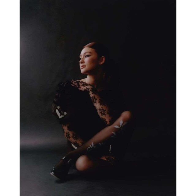 Portrait of a woman with lace gloves and floral-patterned tights, sitting indoors in dark, moody lighting, looking off-camera.