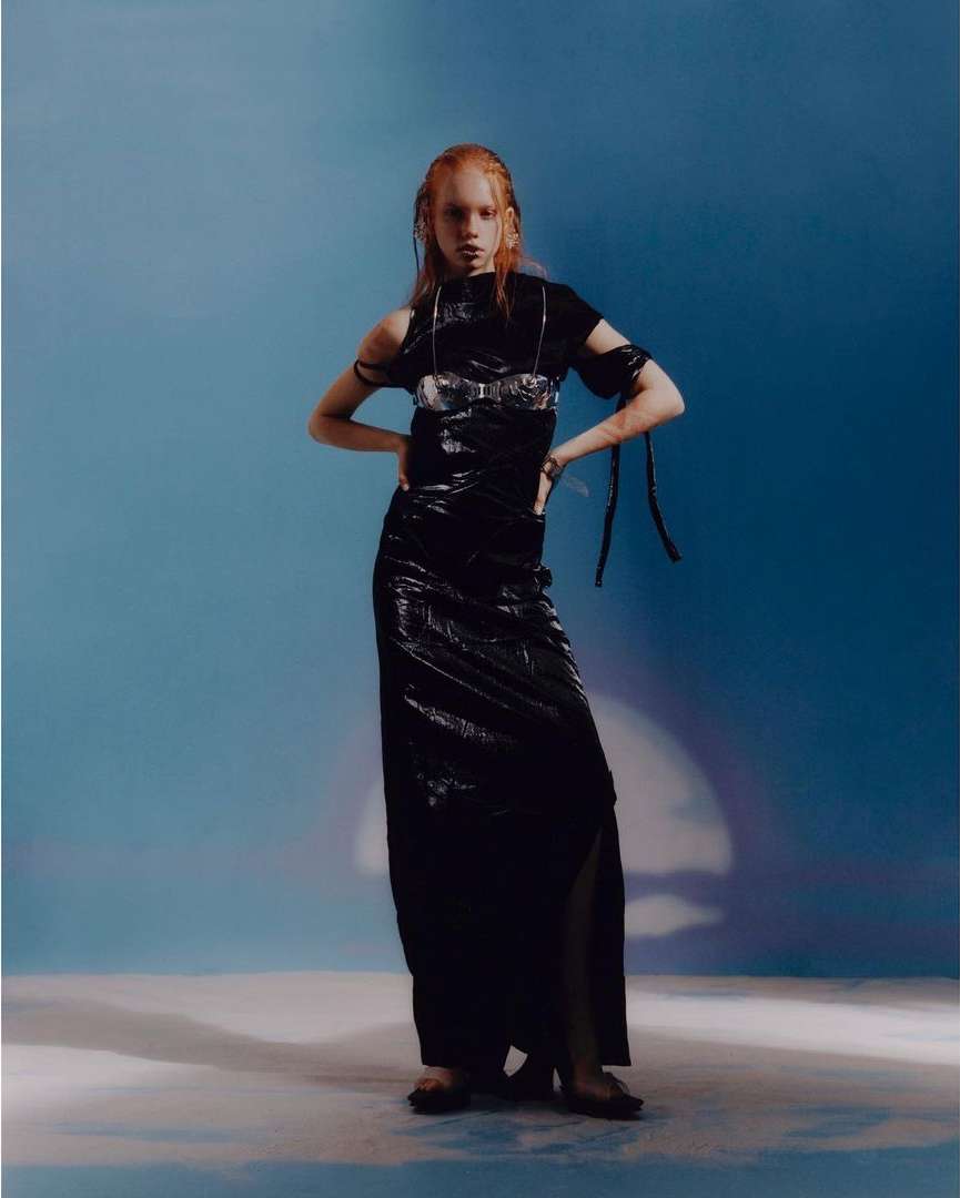 A fair-skinned adult female with red hair poses in a wet-look black dress against a blue gradient background in a studio setting.