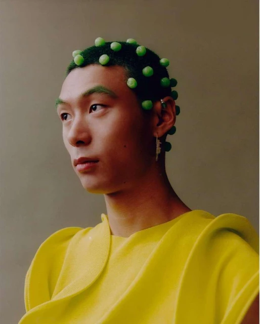 A portrait of an androgynous person with short, dark green hair adorned with round, green baubles, set against a neutral backdrop.