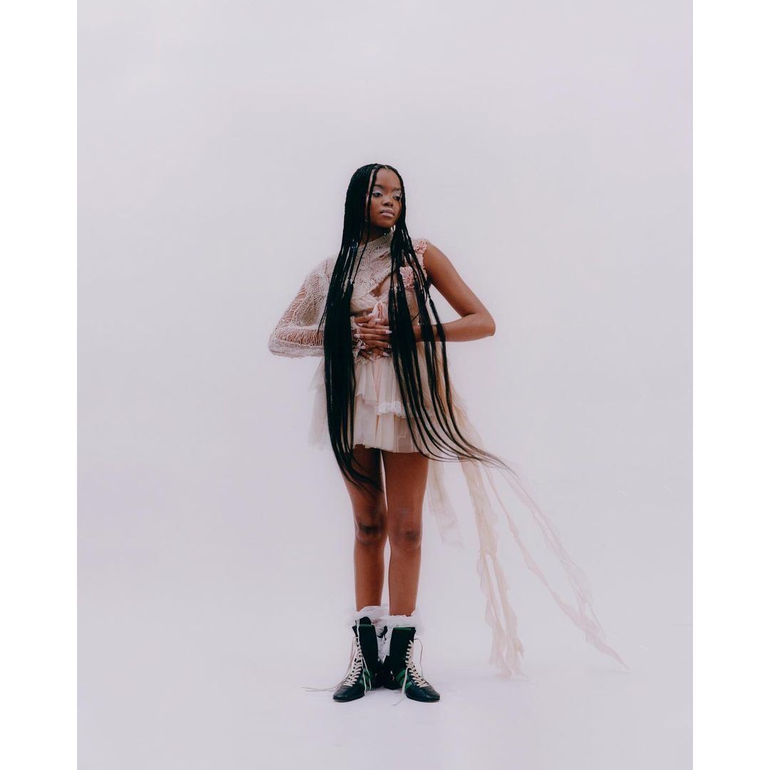 Woman in a lacy dress with long braids stands in a bright studio setting, accented by stylish winter boots, creating a fashion-forward image.