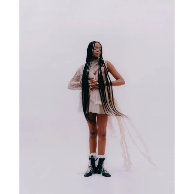 Woman in a lacy dress with long braids stands in a bright studio setting, accented by stylish winter boots, creating a fashion-forward image.