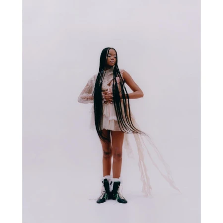 Woman in a lacy dress with long braids stands in a bright studio setting, accented by stylish winter boots, creating a fashion-forward image.