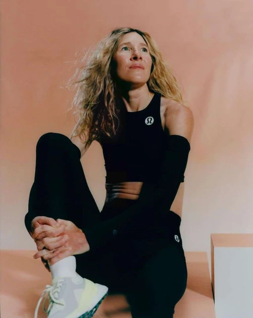 Athletic woman with long blonde hair poses indoors wearing black workout clothes and white sneakers against a peach colored wall.