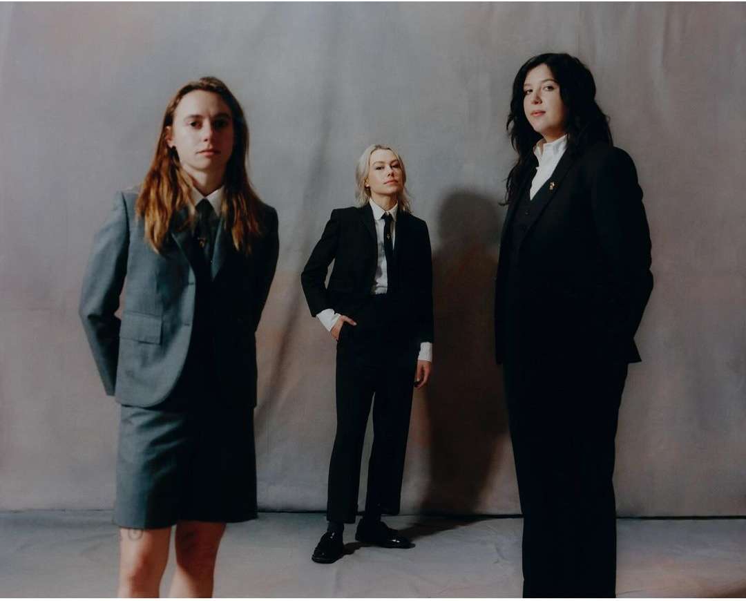 Three women stand in formal suits against a neutral background; one in a grey skirt suit, two in black pantsuits, all looking forward.