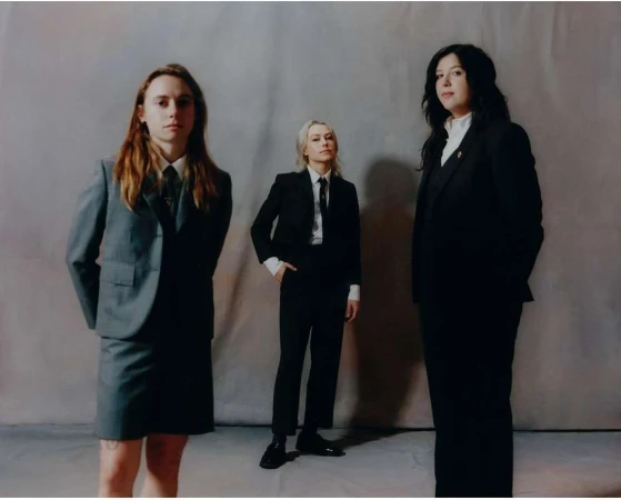 Three women stand in formal suits against a neutral background; one in a grey skirt suit, two in black pantsuits, all looking forward.