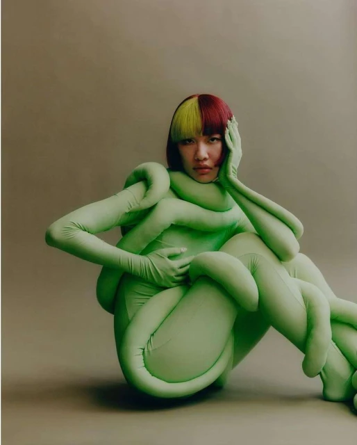 A woman with striking two-tone hair poses in a light green full body suit with padded, tube-like accents, creating a surreal, sculptural look.