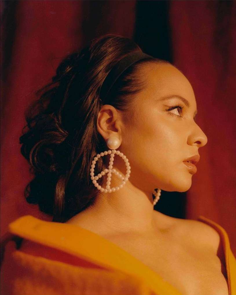 Profile portrait of a woman with pearl peace-sign earrings, wearing an orange top, against a reddish-brown backdrop.