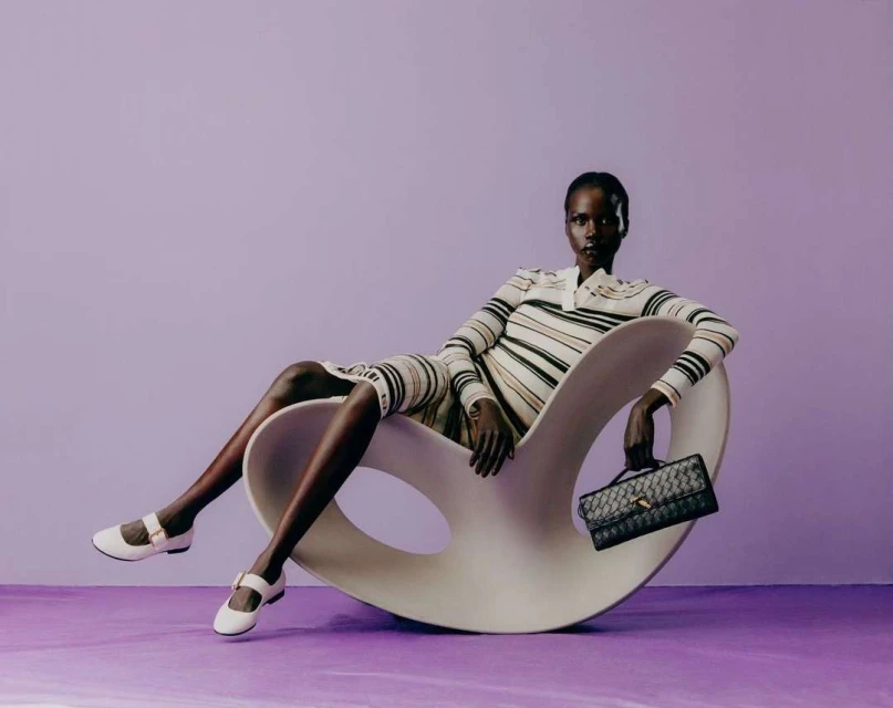 Elegant adult woman posing in a striped long-sleeve dress while sitting in a modern rocking chair and holding a black handbag against a purple backdrop.