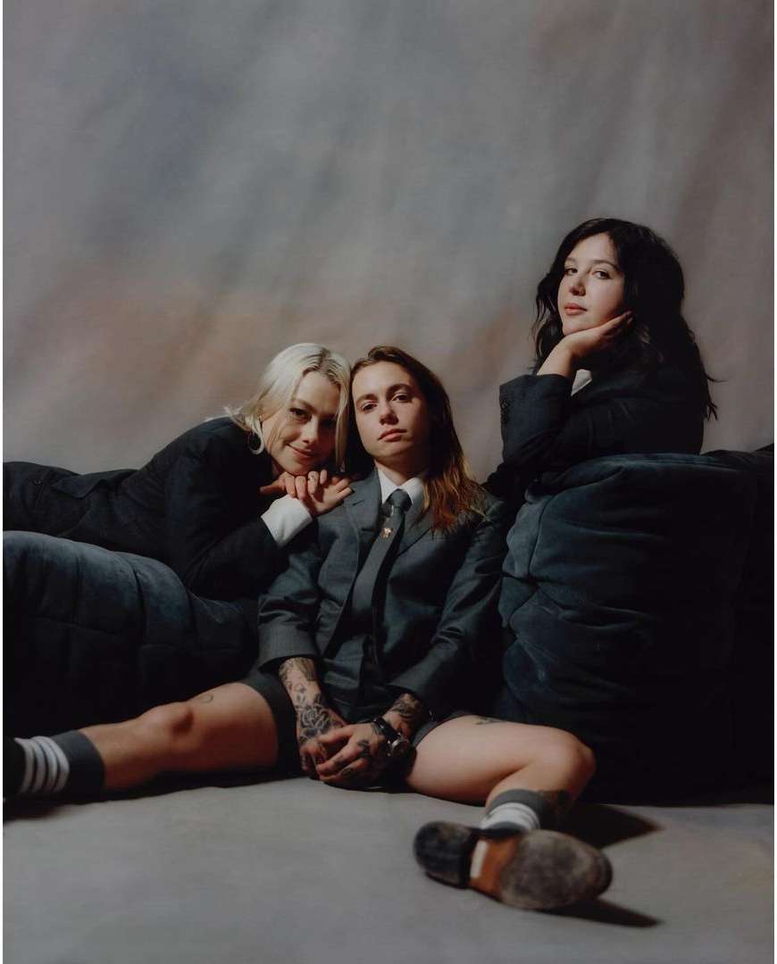 Three women pose in blazers and shorts, seated on a floor with a muted backdrop. One woman wears a tie and patterned socks.