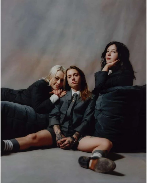 Three women pose in blazers and shorts, seated on a floor with a muted backdrop. One woman wears a tie and patterned socks.