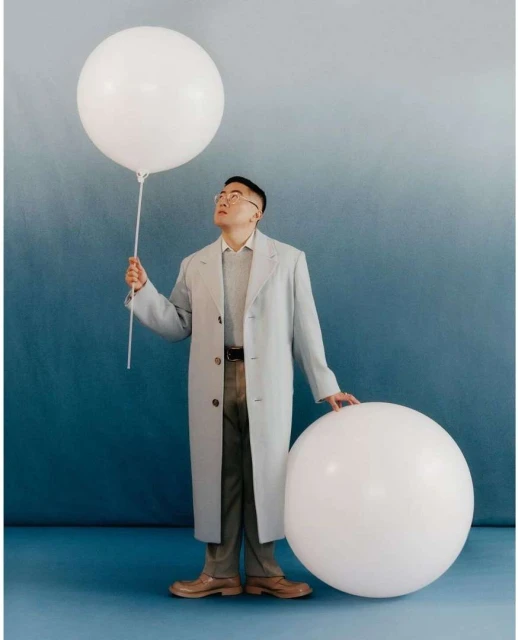 Young man in glasses and coat poses in a studio with large white balloons against a blue gradient backdrop in a medium shot.