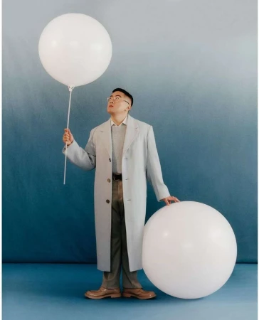 Young man in glasses and coat poses in a studio with large white balloons against a blue gradient backdrop in a medium shot.