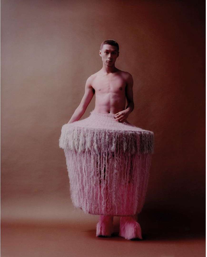 A shirtless young adult male poses in a pink, furry costume against a solid tan backdrop in a studio-lit fashion shot.