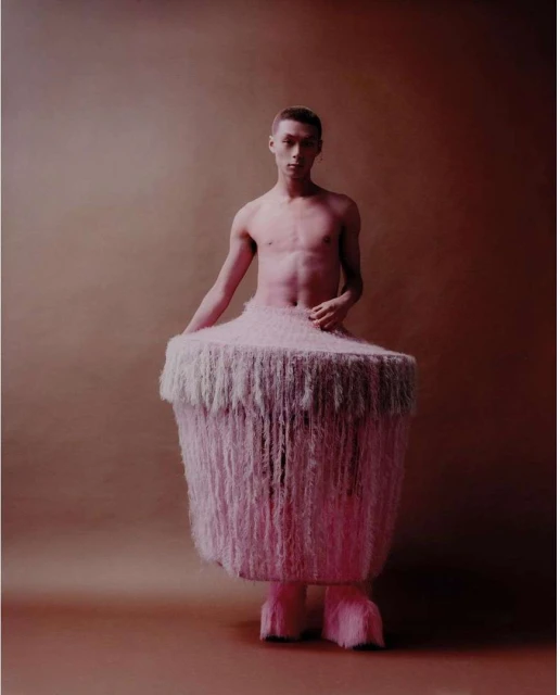 A shirtless young adult male poses in a pink, furry costume against a solid tan backdrop in a studio-lit fashion shot.