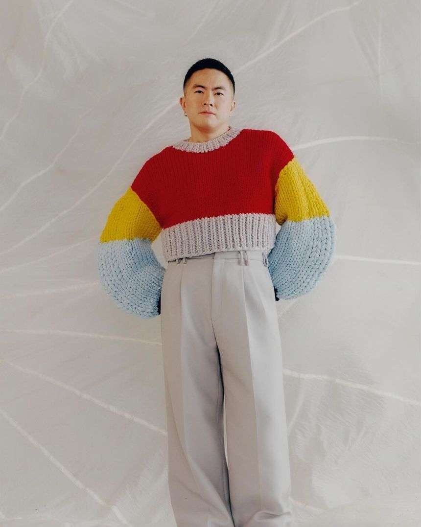 Portrait of a young adult male in a color-block knit sweater and grey trousers posing against a textured white backdrop in a studio setting.